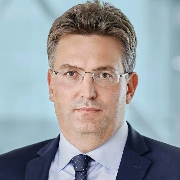 Professional headshot of a man with short brown hair wearing glasses, a dark suit, light blue shirt, and dark tie, against a blurred light blue background.