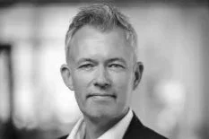 A middle-aged man with short gray hair in a business suit, standing outdoors with a blurred city background.
