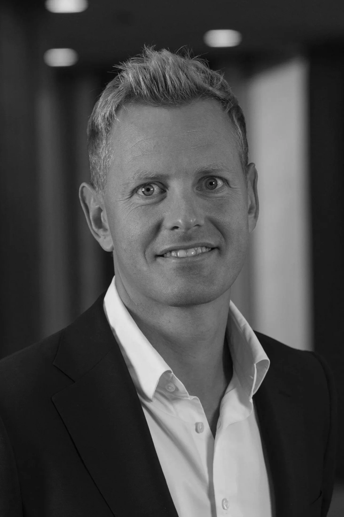 A man with blonde hair and blue eyes wearing a white dress shirt and black suit jacket, standing indoors with a blurred background.