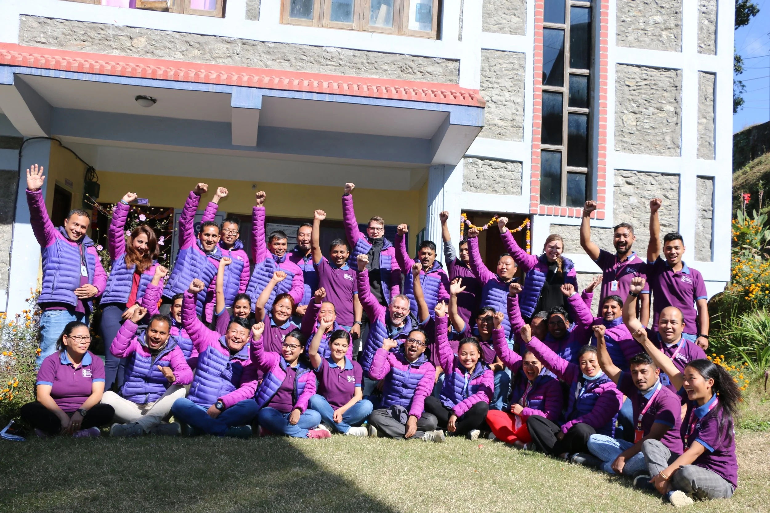 Group of smiling people wearing purple jackets and shirts, celebrating outdoors in front of a building with stone and white trim, some raising fists in the air.