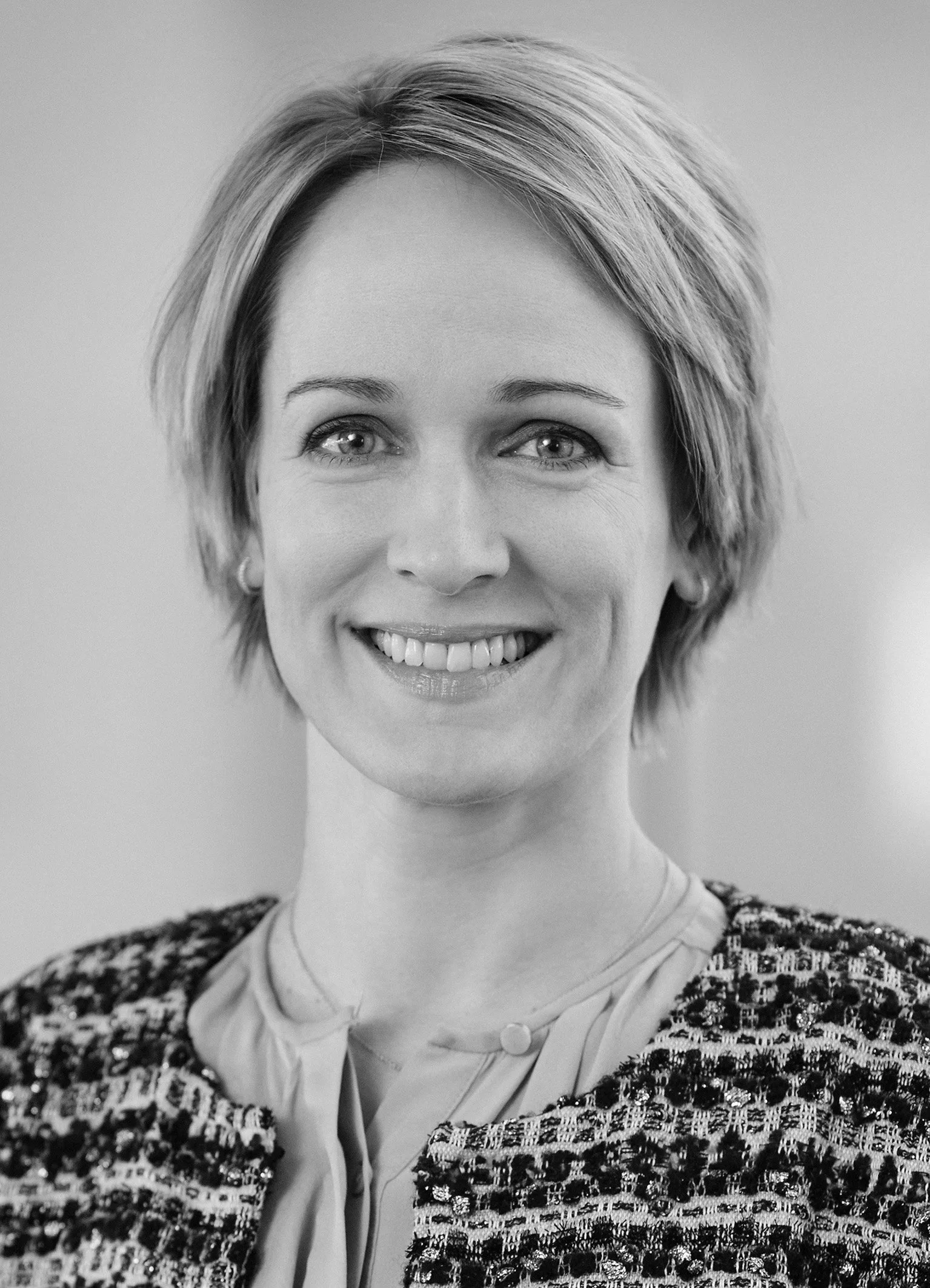 Close-up of a smiling woman with short blonde hair, wearing a tweed jacket and beige blouse, against a neutral background.