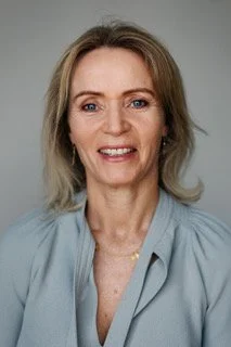 Smiling woman with blonde hair, wearing a light blue blouse, facing forward against a plain background.