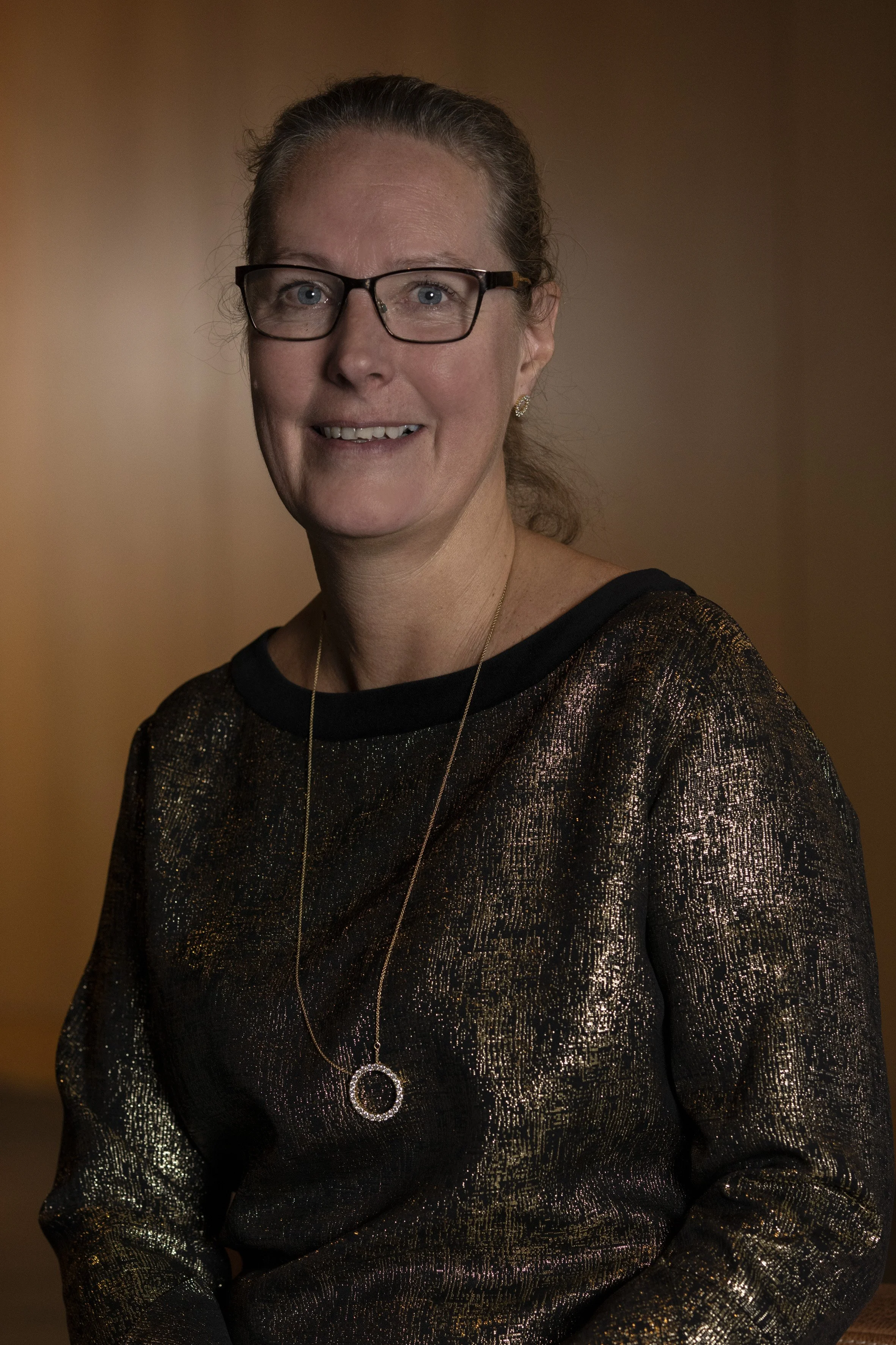 A middle-aged woman with glasses, wearing a black and gold patterned top and a necklace with a circular pendant, smiling at the camera.