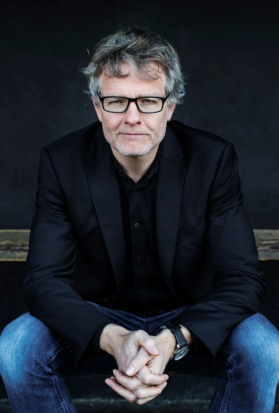 A middle-aged man with gray hair and glasses, wearing a black blazer and black shirt, sitting with hands clasped, against a dark background.