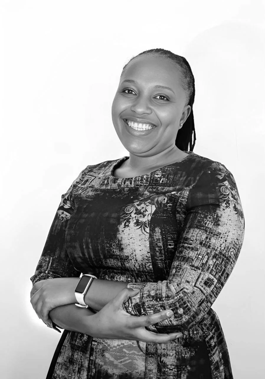 A smiling woman with braided hair, cross-armed, wearing a patterned dress and a smartwatch, standing against a white background.