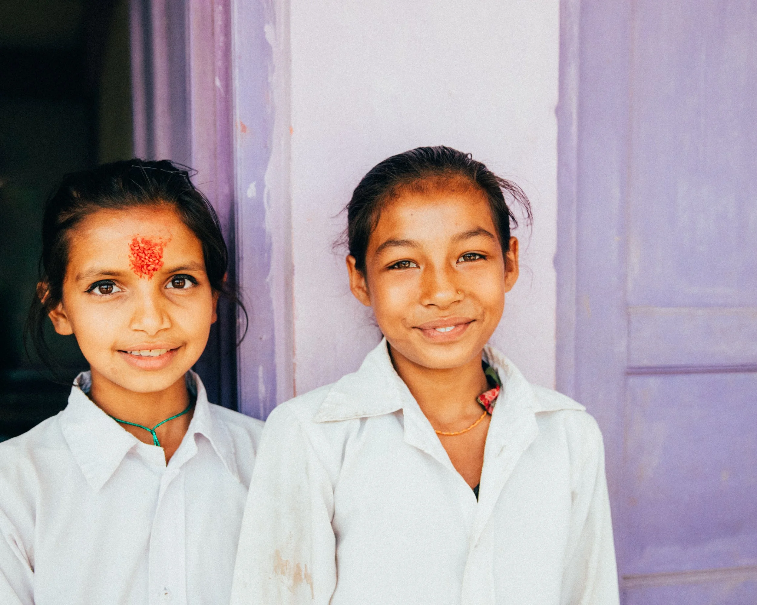 Two young girls in white shirts standing in front of a purple wall, smiling at the camera.