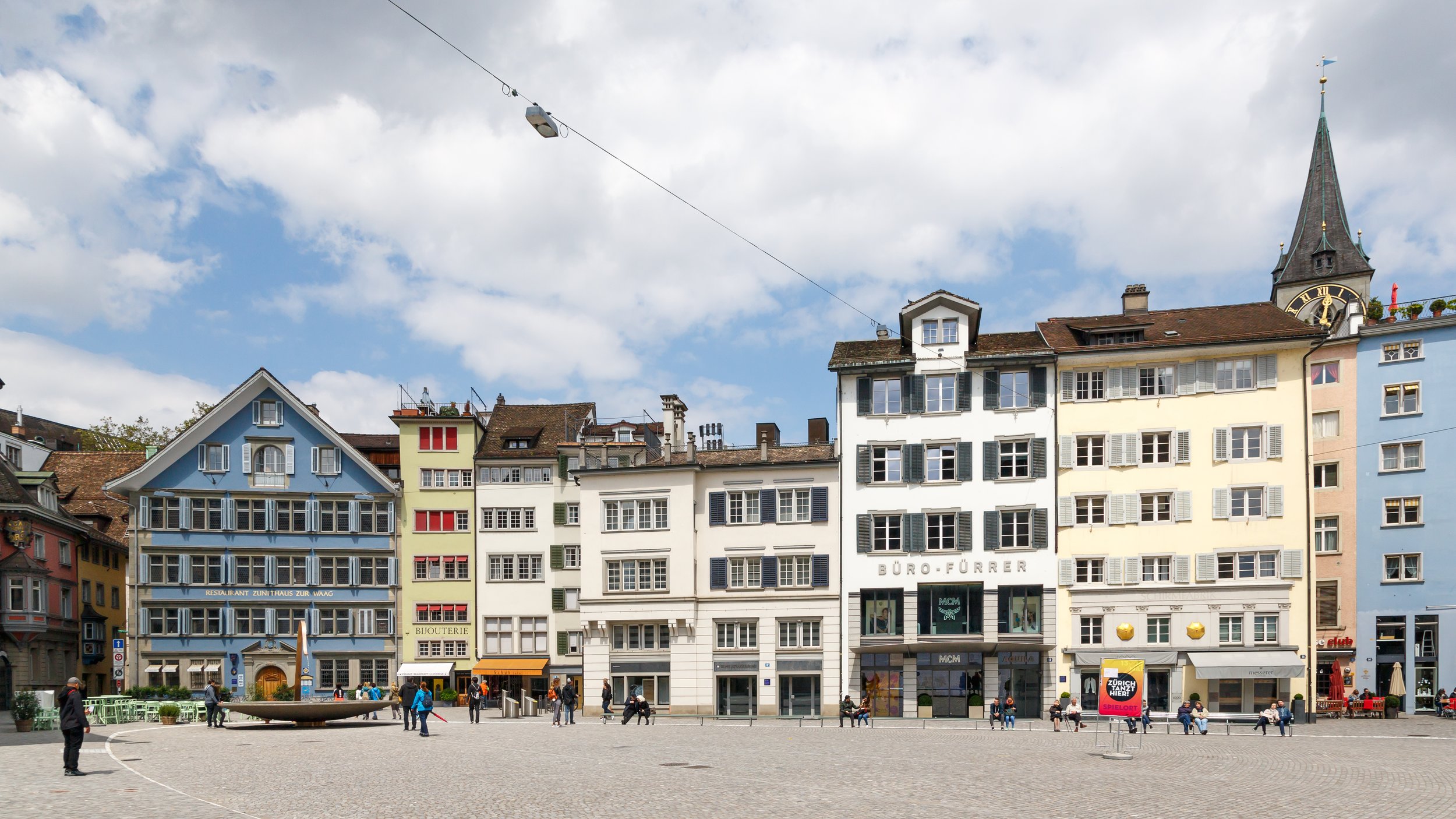 A European city square with colorful, multi-story buildings, some with shops on the ground floor, and people walking and sitting on benches. A church steeple is visible in the background.