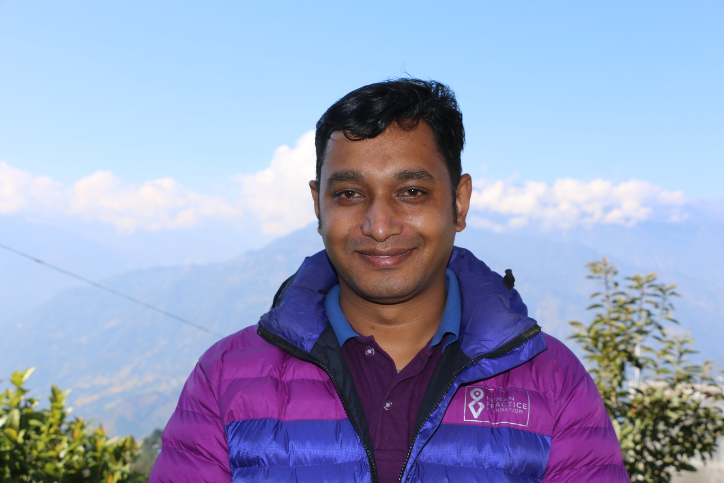 A man smiling outdoors in front of mountains and blue sky, wearing a purple and blue jacket with a logo on the chest.
