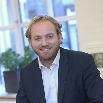 A man with blond hair and a beard, wearing a dark suit jacket and white shirt, smiling indoors with large windows and potted plants in the background.