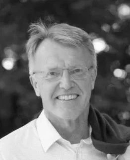 A smiling middle-aged man wearing glasses and a white button-up shirt, holding a navy jacket over his shoulder, standing outdoors in a park with green trees and grass in the background.