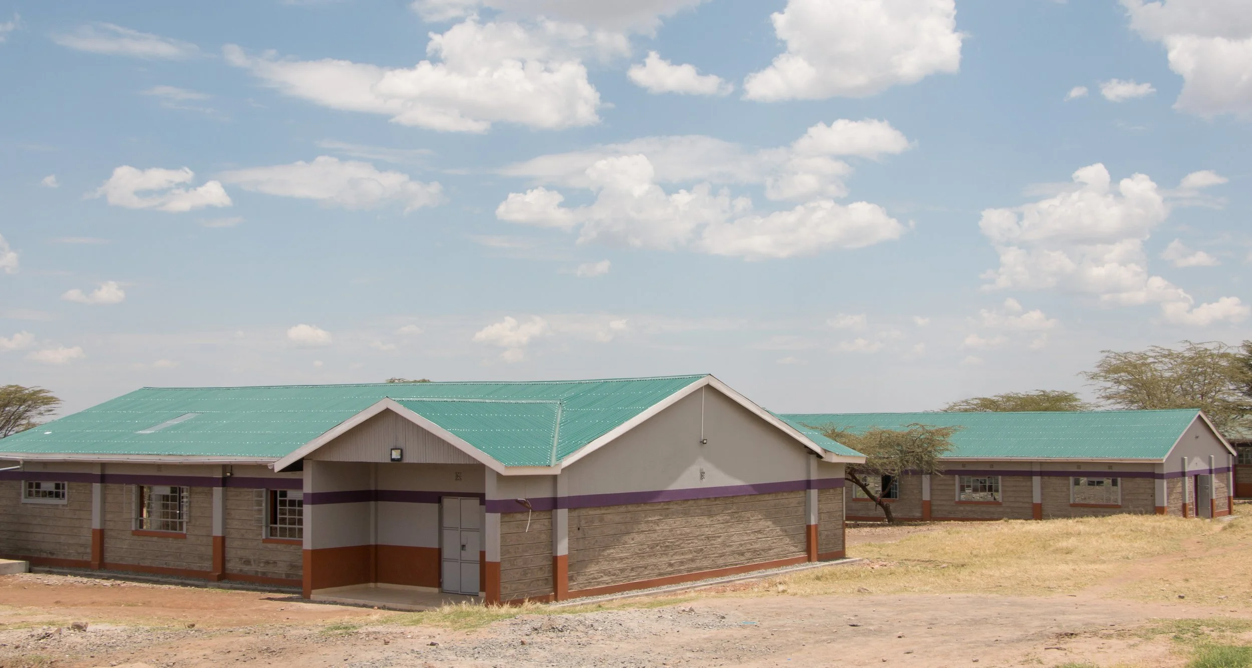 A single-story building with a green metal roof and beige walls, featuring painted stripes, in a dry, open landscape under a blue sky with scattered clouds.