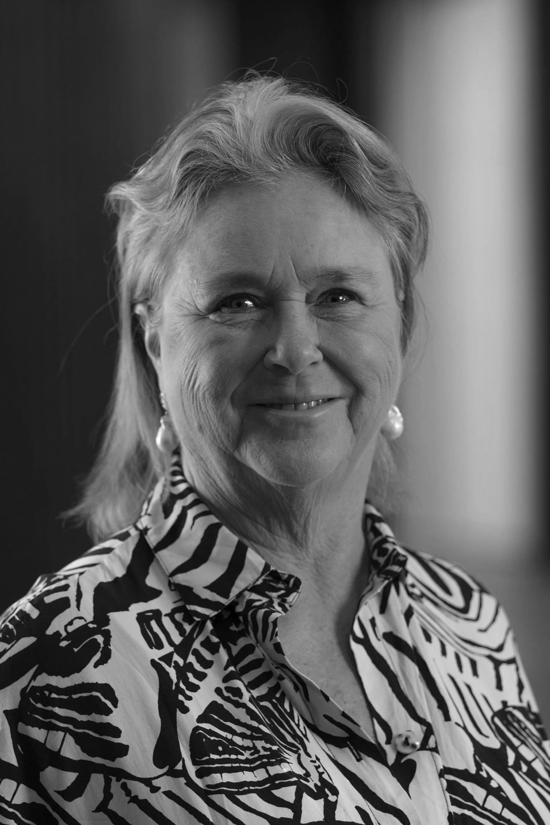 An older woman with blonde hair, wearing a black and white patterned blouse with butterfly and zebra designs, smiling with pearl earrings, standing indoors.