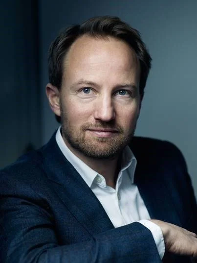 A man with light skin and short, dark hair, wearing a white shirt and dark blazer, looking slightly to the side in front of a dark, plain background.