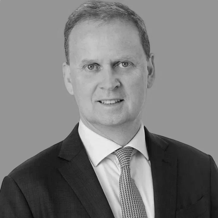 A professional headshot of a man wearing a dark suit, light blue shirt, and patterned tie against a plain grey background.