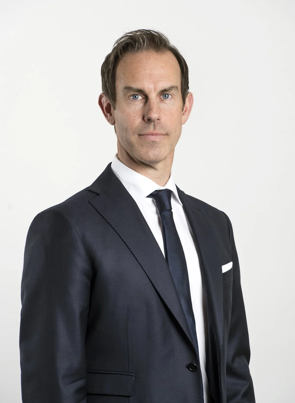 A man with light skin, brown hair, and blue eyes wearing a dark suit, white shirt, and dark tie against a plain white background.