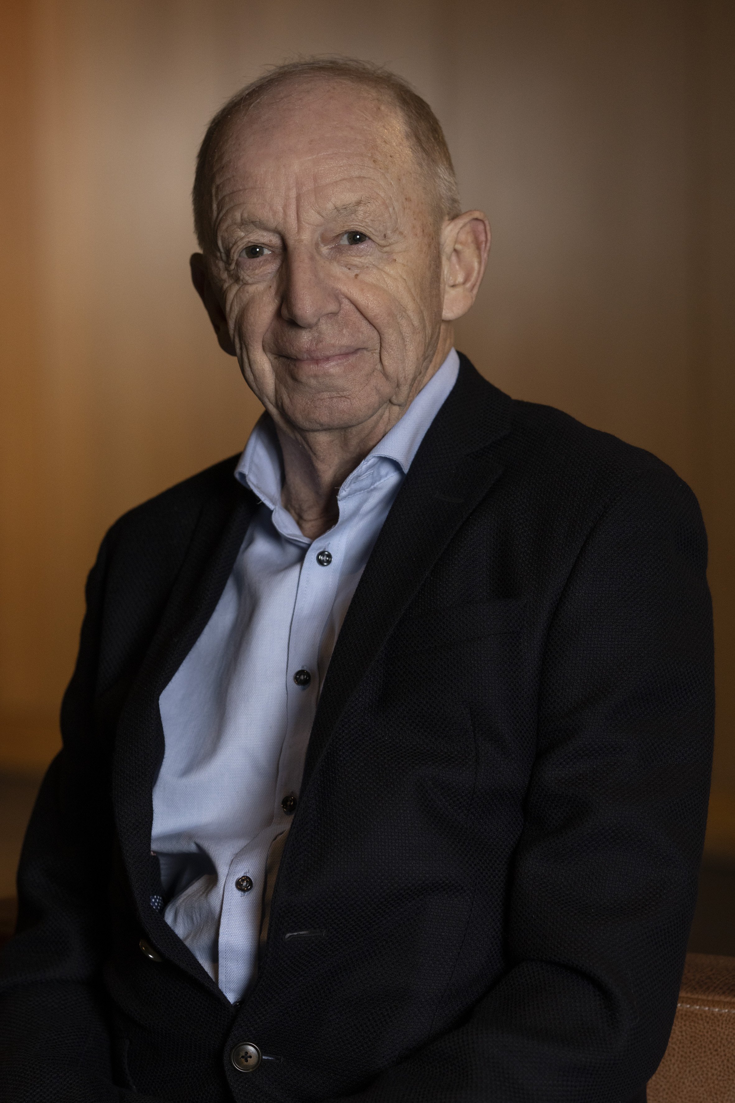 An older man with a gentle smile, wearing a light blue dress shirt and a dark blazer, seated in a softly lit room.