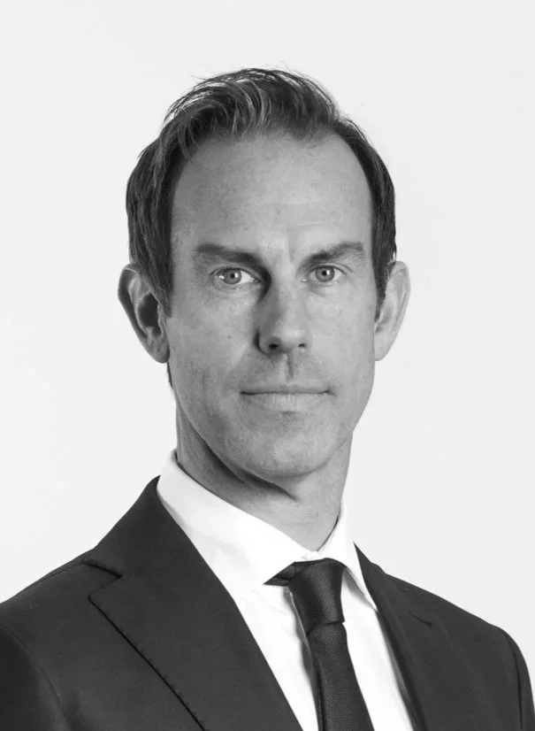 A man with light skin, brown hair, and blue eyes wearing a dark suit, white shirt, and dark tie against a plain white background.