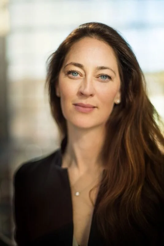A woman with long brown hair and blue eyes posing for a portrait indoors with a blurred background.