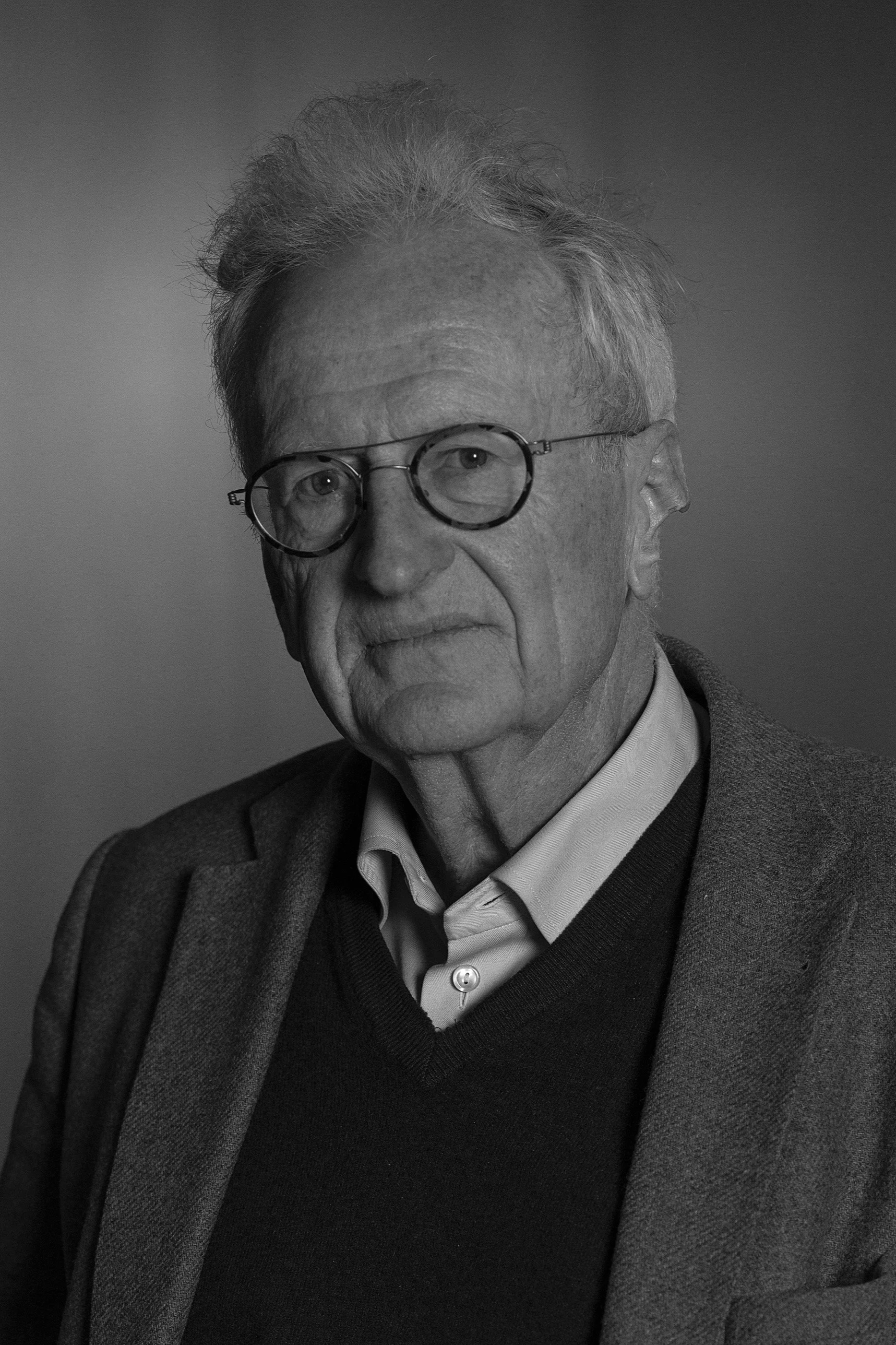 An elderly man with gray hair and glasses, wearing a blue shirt, black sweater, and brown blazer, sitting and looking at the camera against a warm, blurred background.