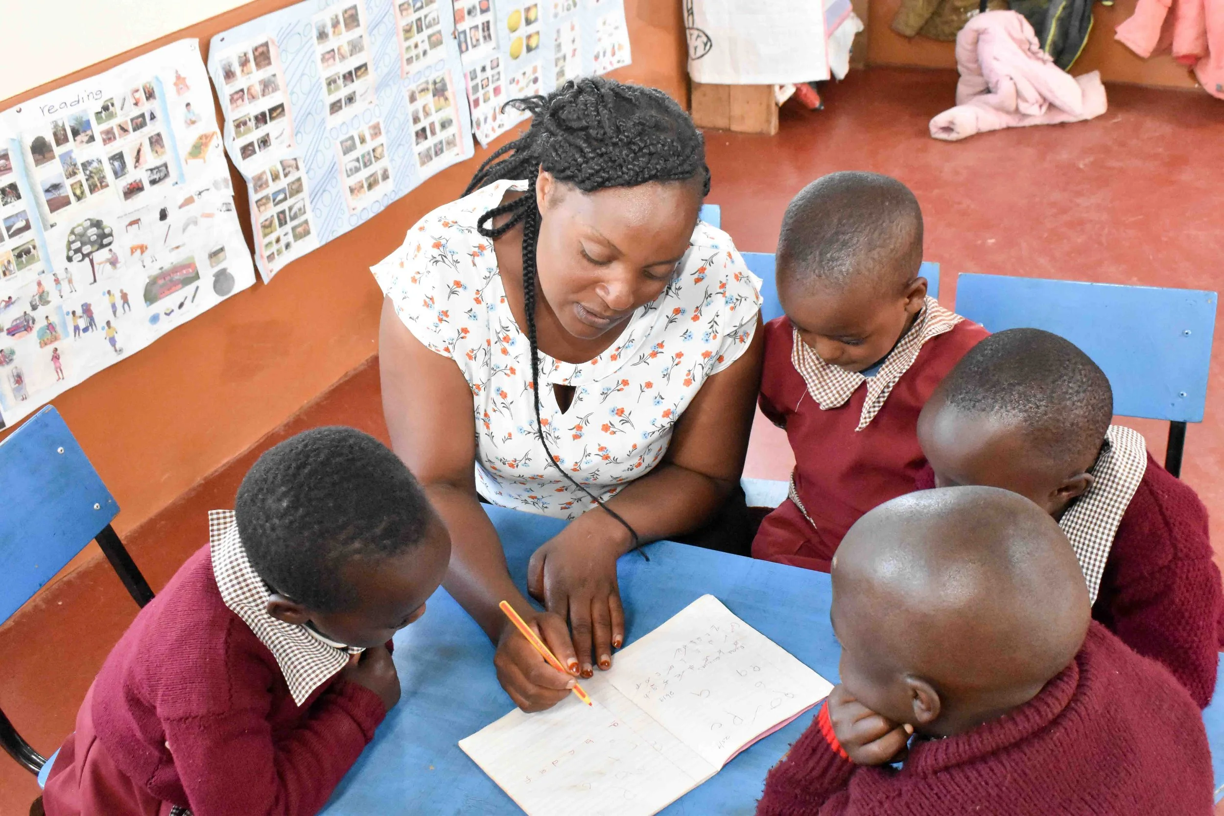 A teacher helping four students with their work at a table in a classroom. The teacher is writing in a notebook, and the students are looking at her and the notebook. The classroom has colorful posters on the wall.