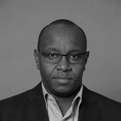 A man in a dark suit jacket and glasses staring at the camera, with a neutral expression, in front of a plain gray background.