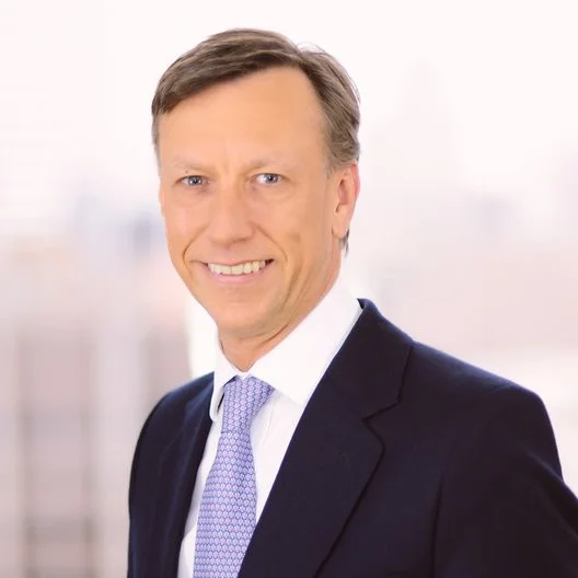 Portrait of a man in a dark suit with a light purple tie, smiling against a blurred office background.