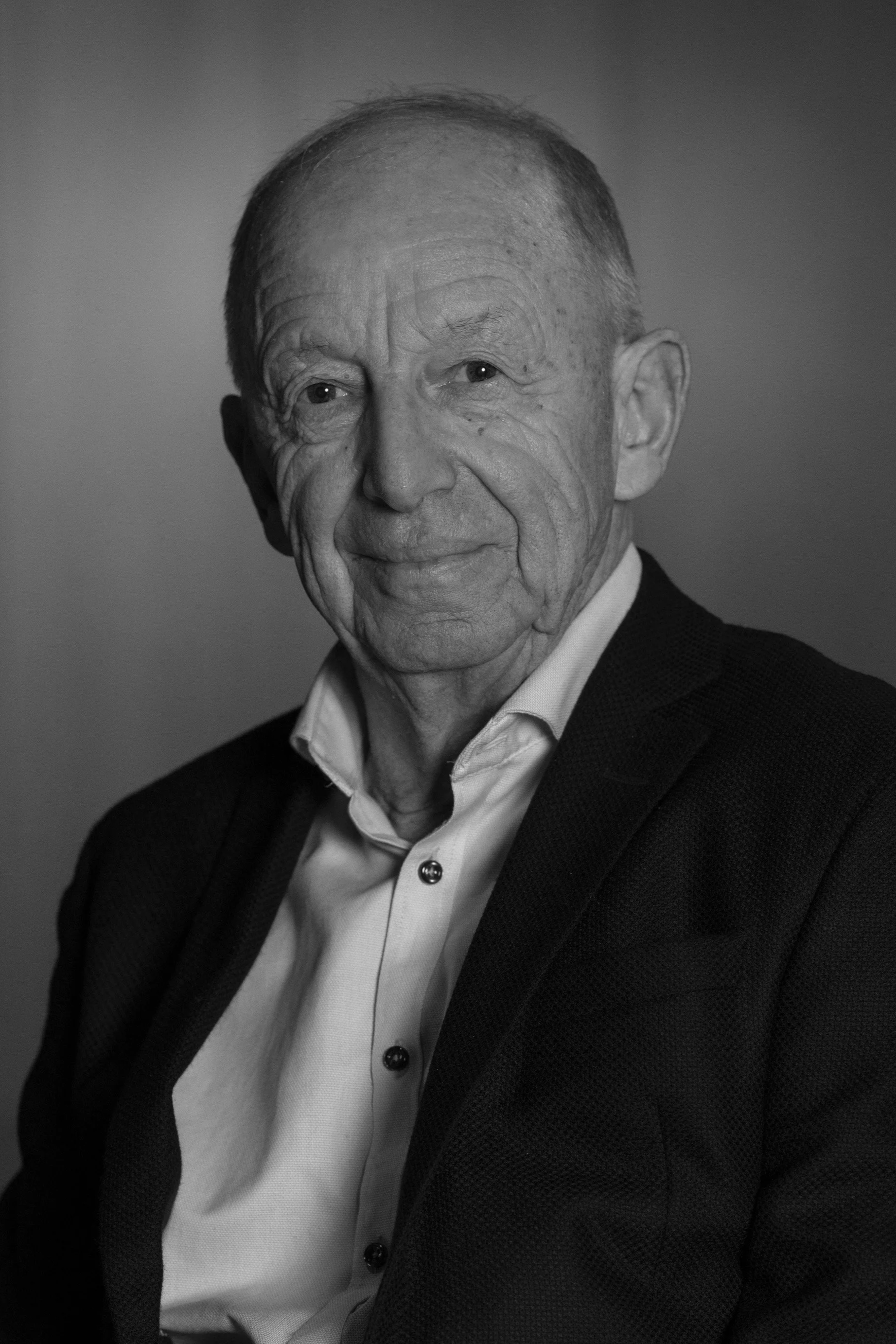 An older man with a gentle smile, wearing a light blue dress shirt and a dark blazer, seated in a softly lit room.
