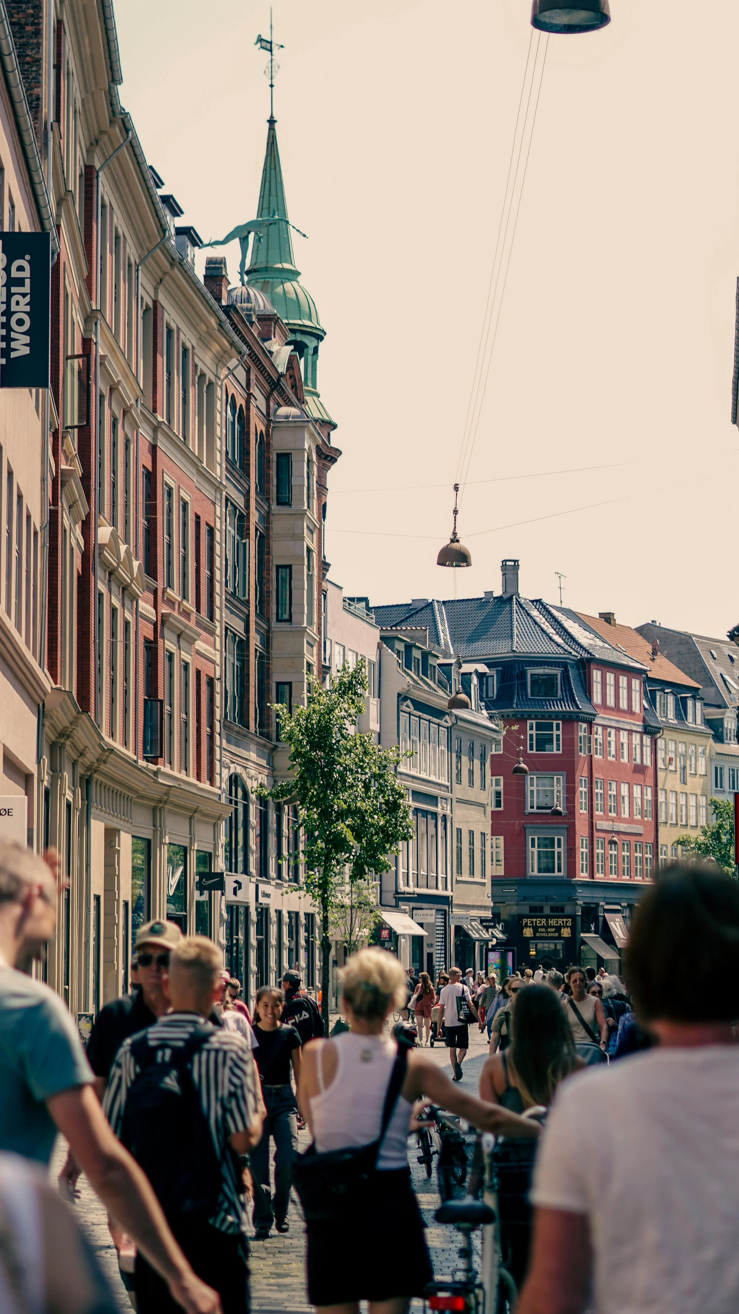 A lively street scene in a European city with colorful old buildings, people walking and biking, and a church steeple in the background.