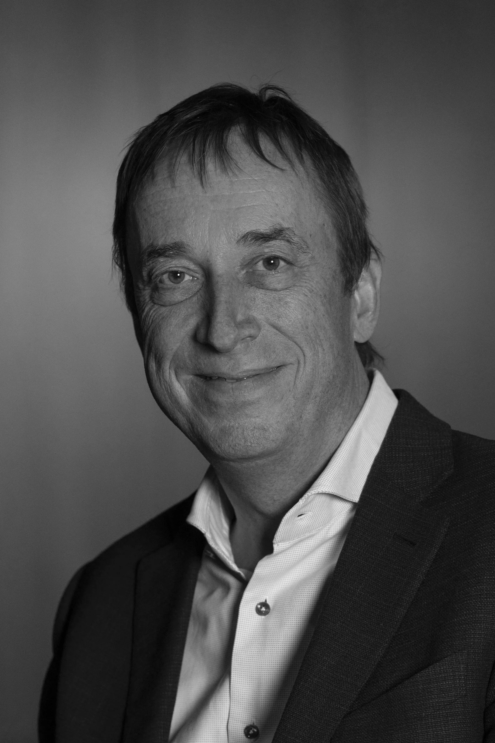 A middle-aged man with short brown hair, wearing a dark blazer and white shirt, sitting against a brown wall.