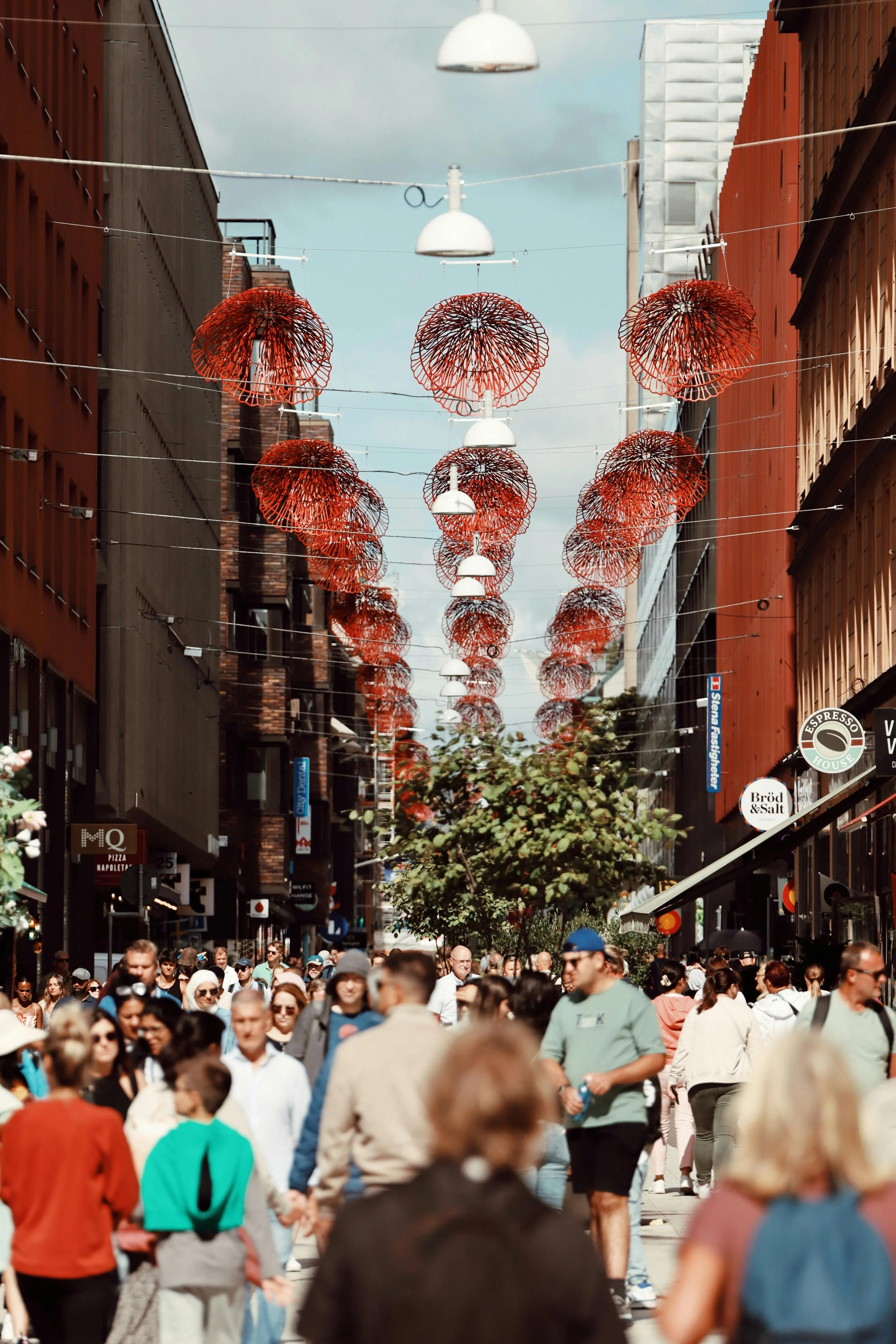 Crowded city street decorated with red and white umbrellas hanging overhead, lined with shops and people walking.