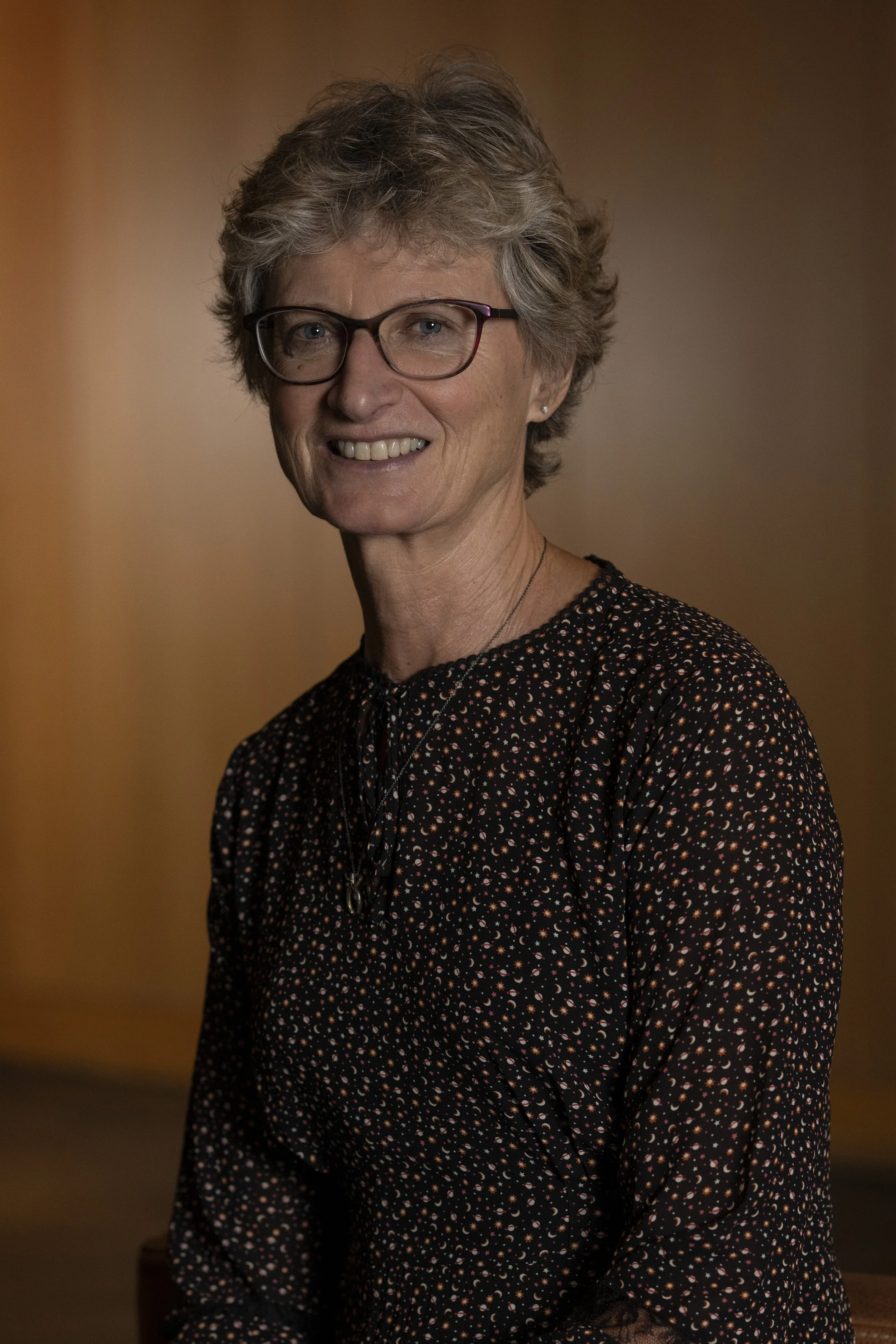 A woman with short gray hair, wearing glasses and a black blouse with small floral patterns, smiling at the camera against a warm brown background.