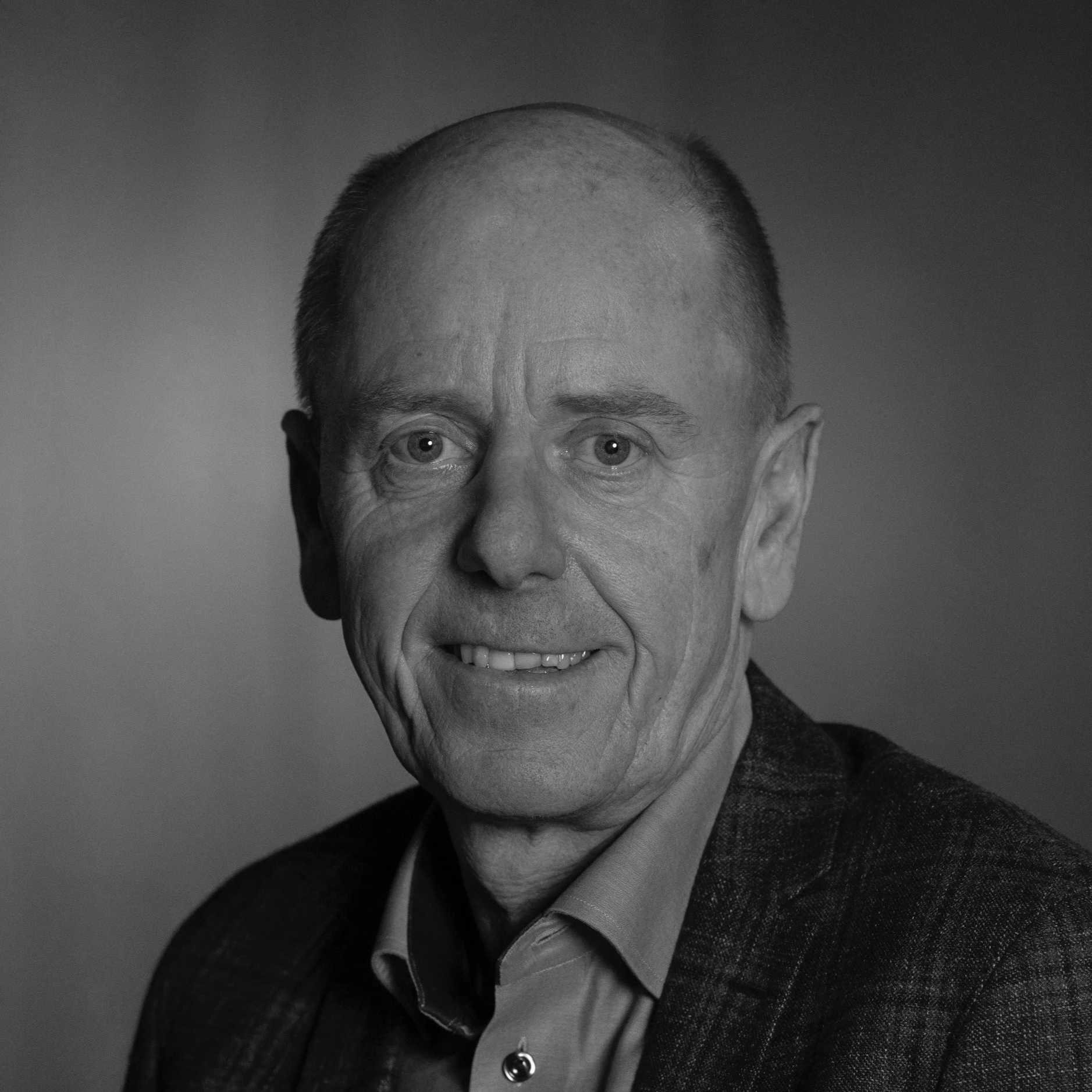 Headshot of an older man with short hair, blue eyes, smiling, wearing a blue button-up shirt and a dark blazer.
