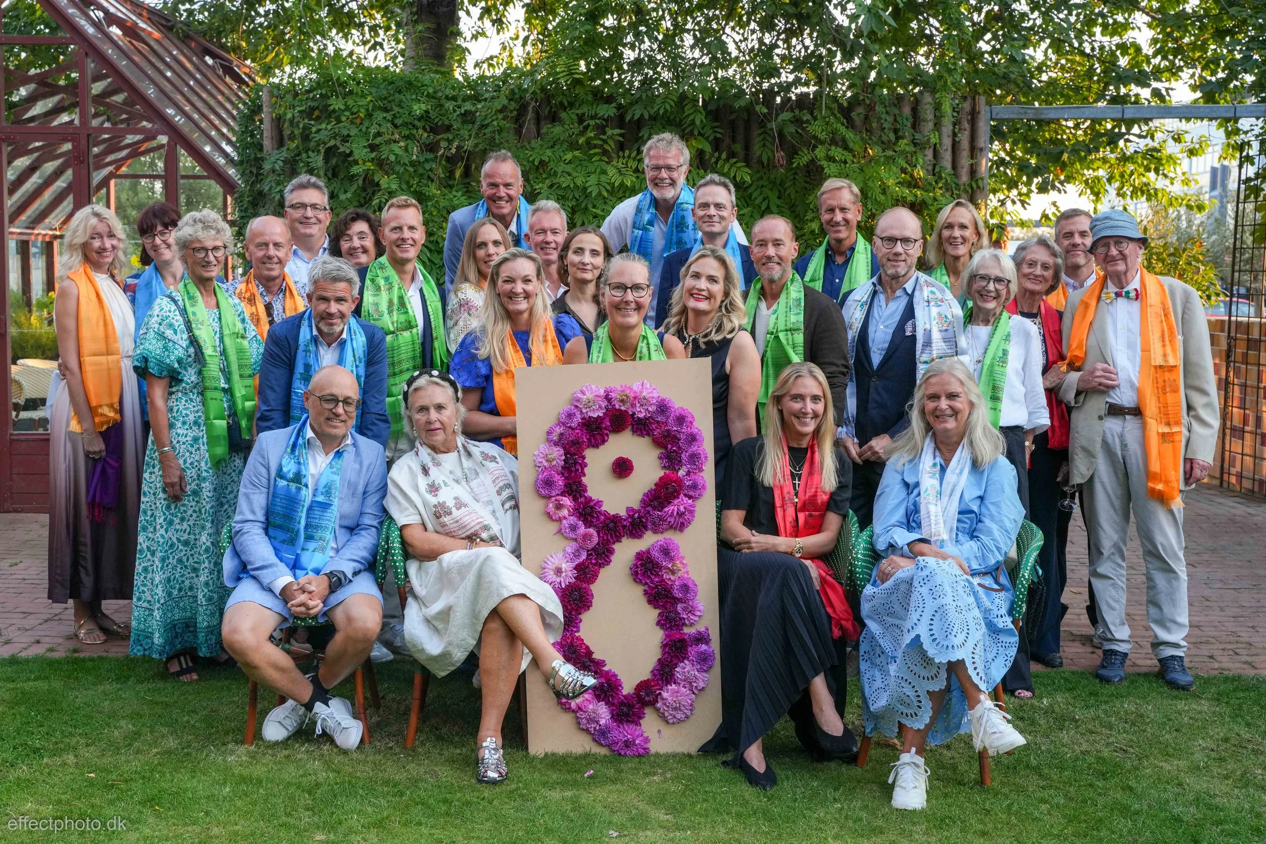 A group of people gathered outdoors for a celebration, with some wearing colorful scarves, and a large floral number nine centerpiece in front of them.