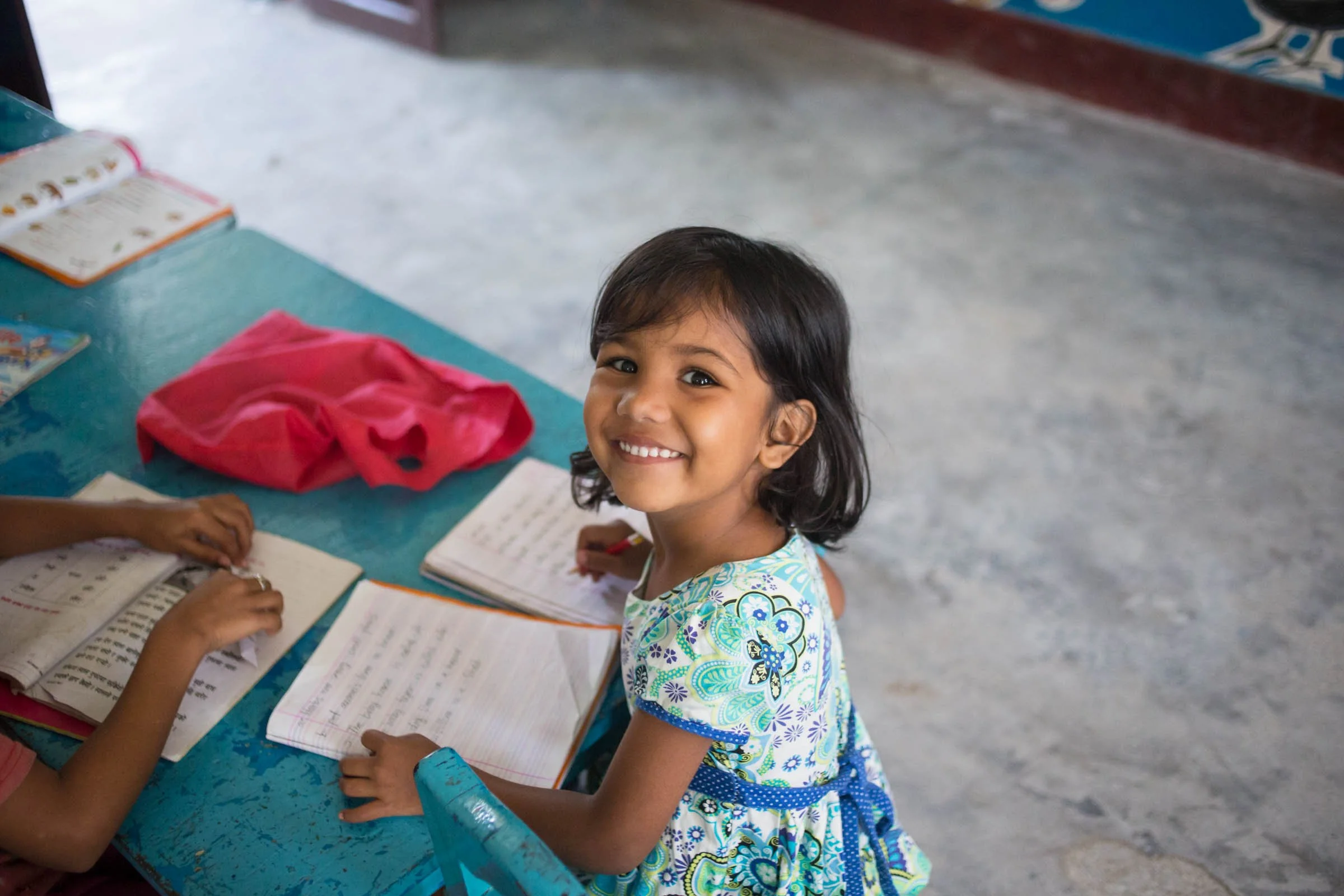 A young girl with short black hair and a colorful dress smiling at the camera while sitting at a blue table with open notebooks and a pink bag.
