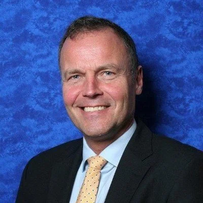 A professional headshot of a smiling man in a suit and tie against a textured blue background.