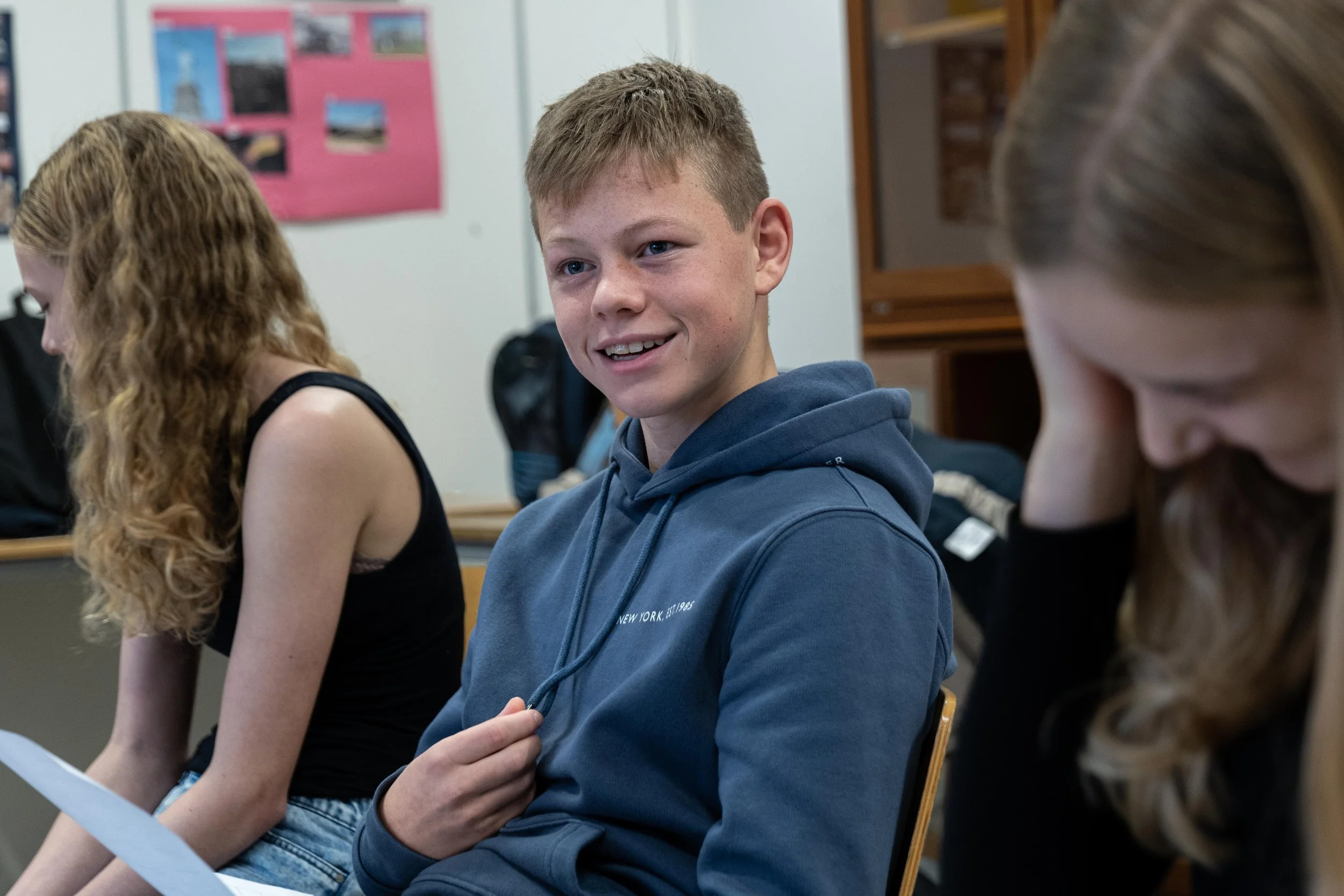 A young boy with short light brown hair, wearing a gray hoodie, sitting in a classroom, smiling and holding the drawstrings of his hoodie. Two girls with long hair, one with red hair in a black tank top and the other with brown hair, are sitting beside him, one looking down and the other looking away.