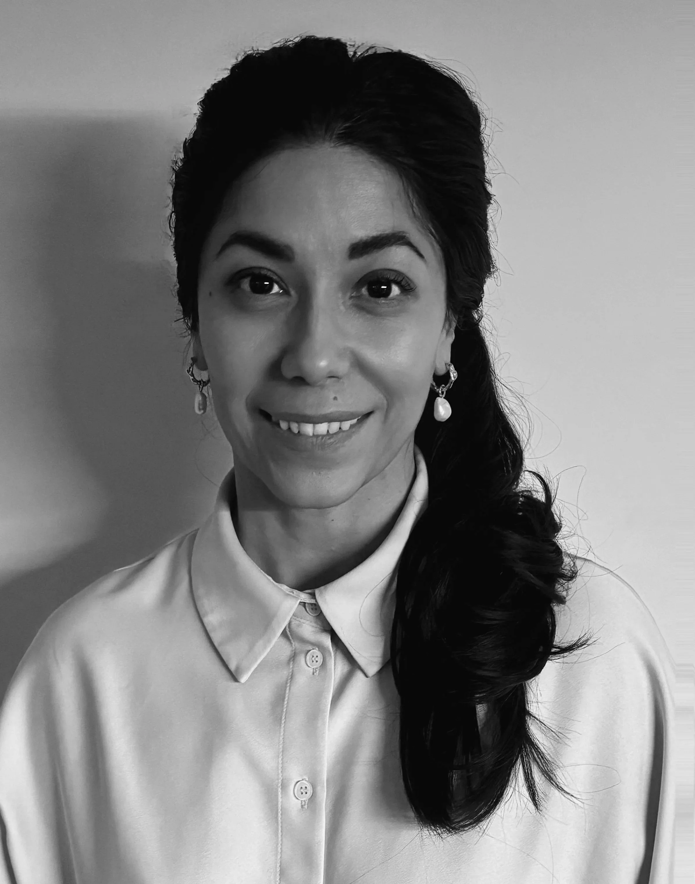 A woman with dark hair styled in loose curls, wearing a cream-colored blouse and pearl earrings, smiling at the camera against a plain light-colored wall.