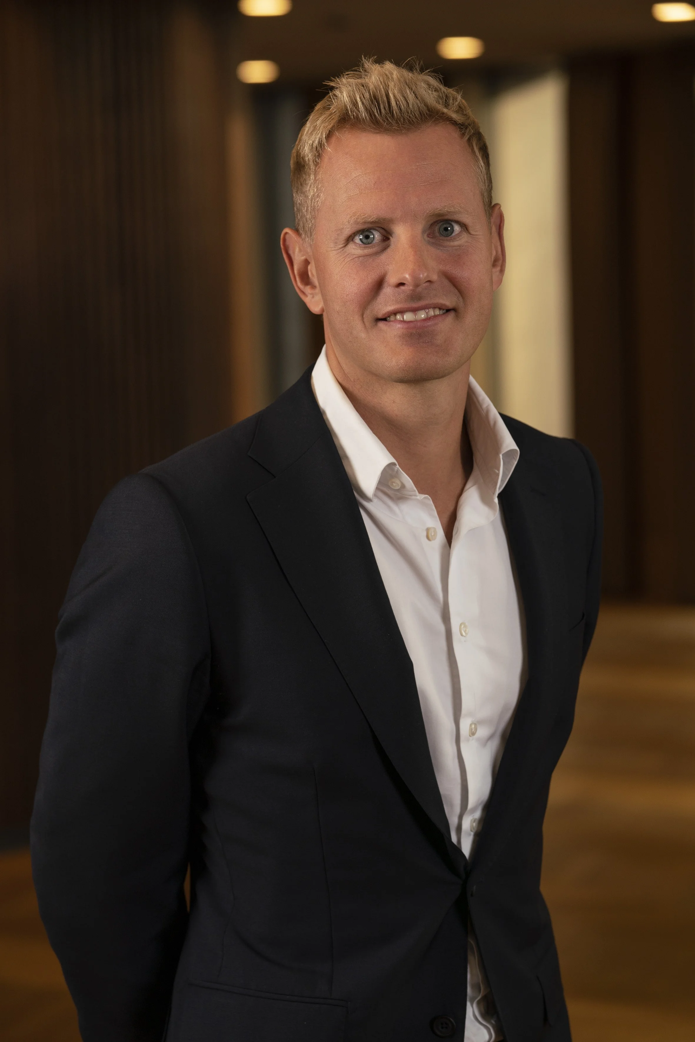 A man with blonde hair and blue eyes wearing a white dress shirt and black suit jacket, standing indoors with a blurred background.