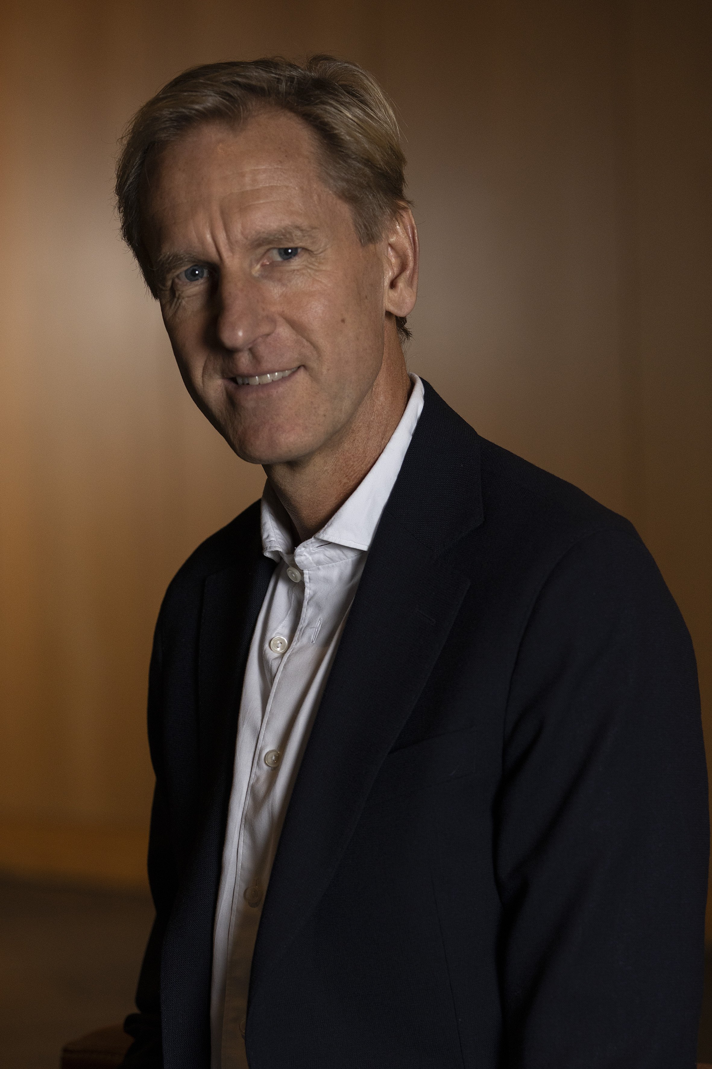 A middle-aged man with blonde hair, wearing a black suit jacket and a white dress shirt, smiling slightly against a warm-colored background.