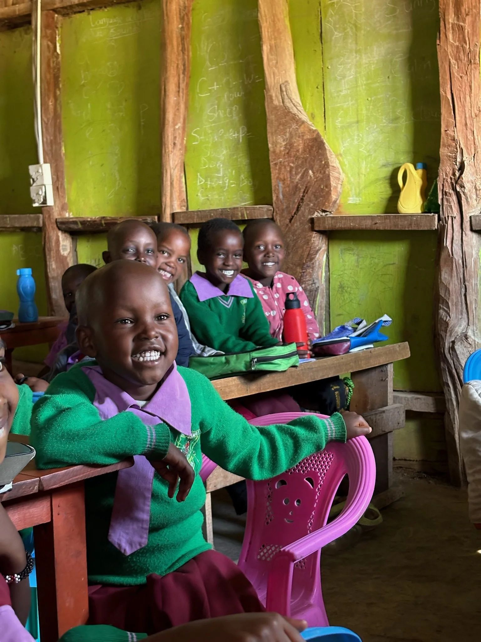 Group of smiling children sitting inside a classroom with wooden walls and green-painted sections, some with water bottles on their desks.