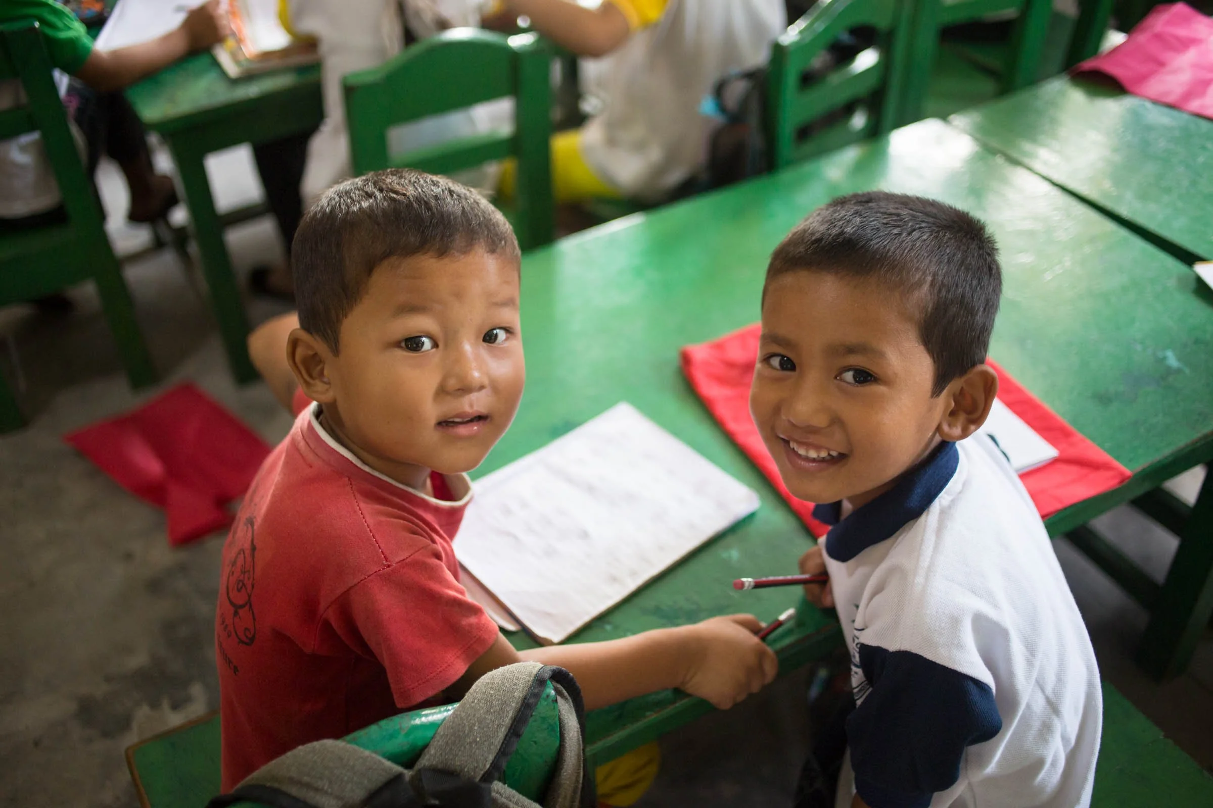 Two young boys sitting at a green school desk, looking up at the camera, with notebooks and pencils in front of them in a classroom with other children in the background.