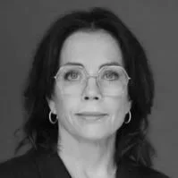 A woman with shoulder-length dark hair, glasses, and hoop earrings, wearing a dark top, against a dark blue background.