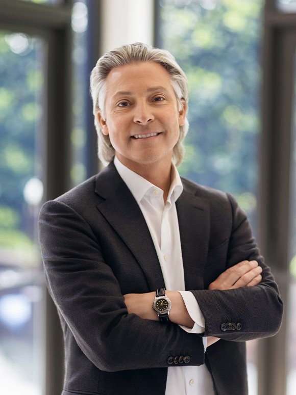 Smiling older man with gray hair, wearing a dark suit and white shirt, crossing his arms, standing indoors with large windows and greenery outside.
