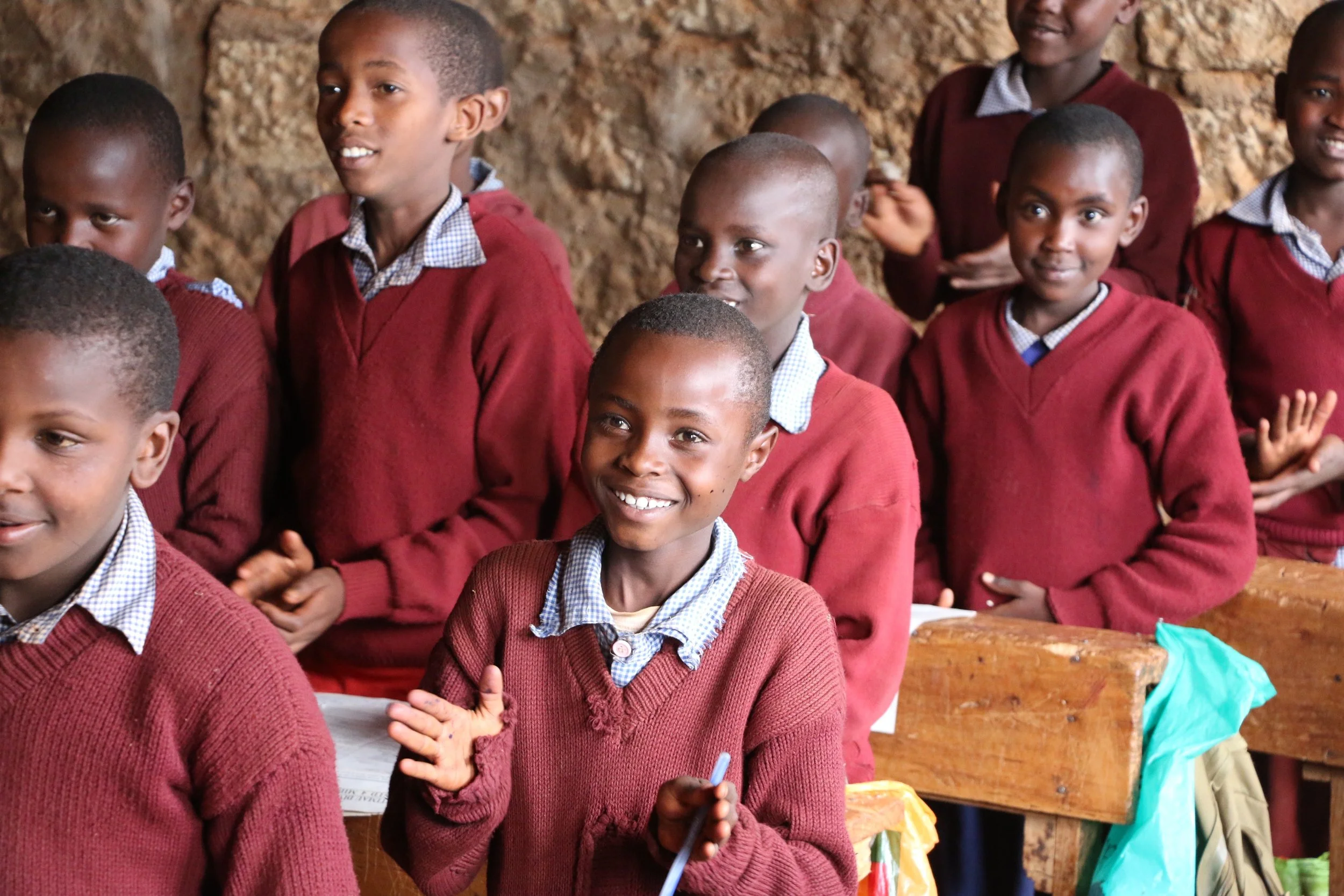 A group of school children in maroon sweaters and checkered shirts in a classroom, some smiling and others clapping, with a stone wall in the background.