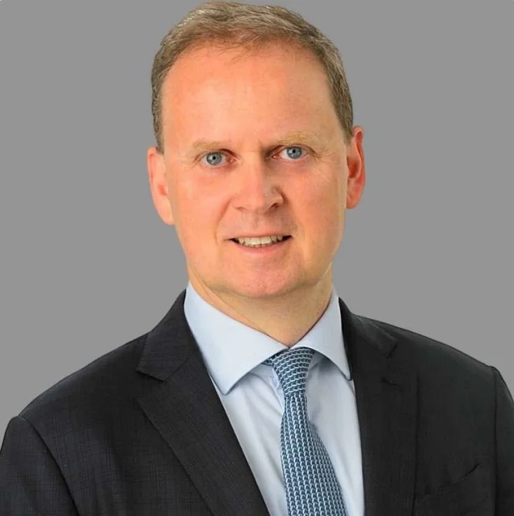A professional headshot of a man wearing a dark suit, light blue shirt, and patterned tie against a plain grey background.