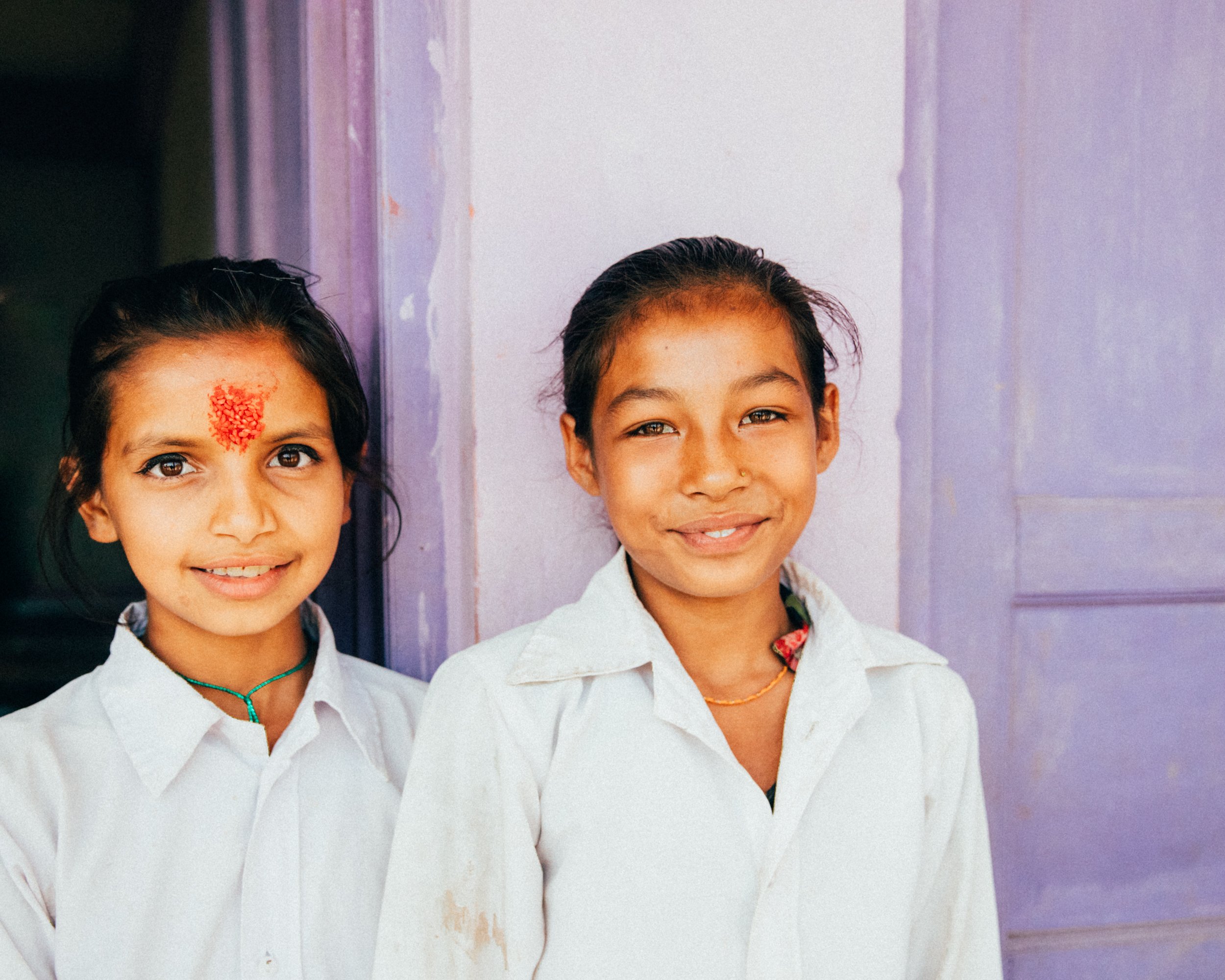 Two smiling girls in white school uniforms standing in front of a purple wall, one with a red mark on her forehead and the other with a necklace.