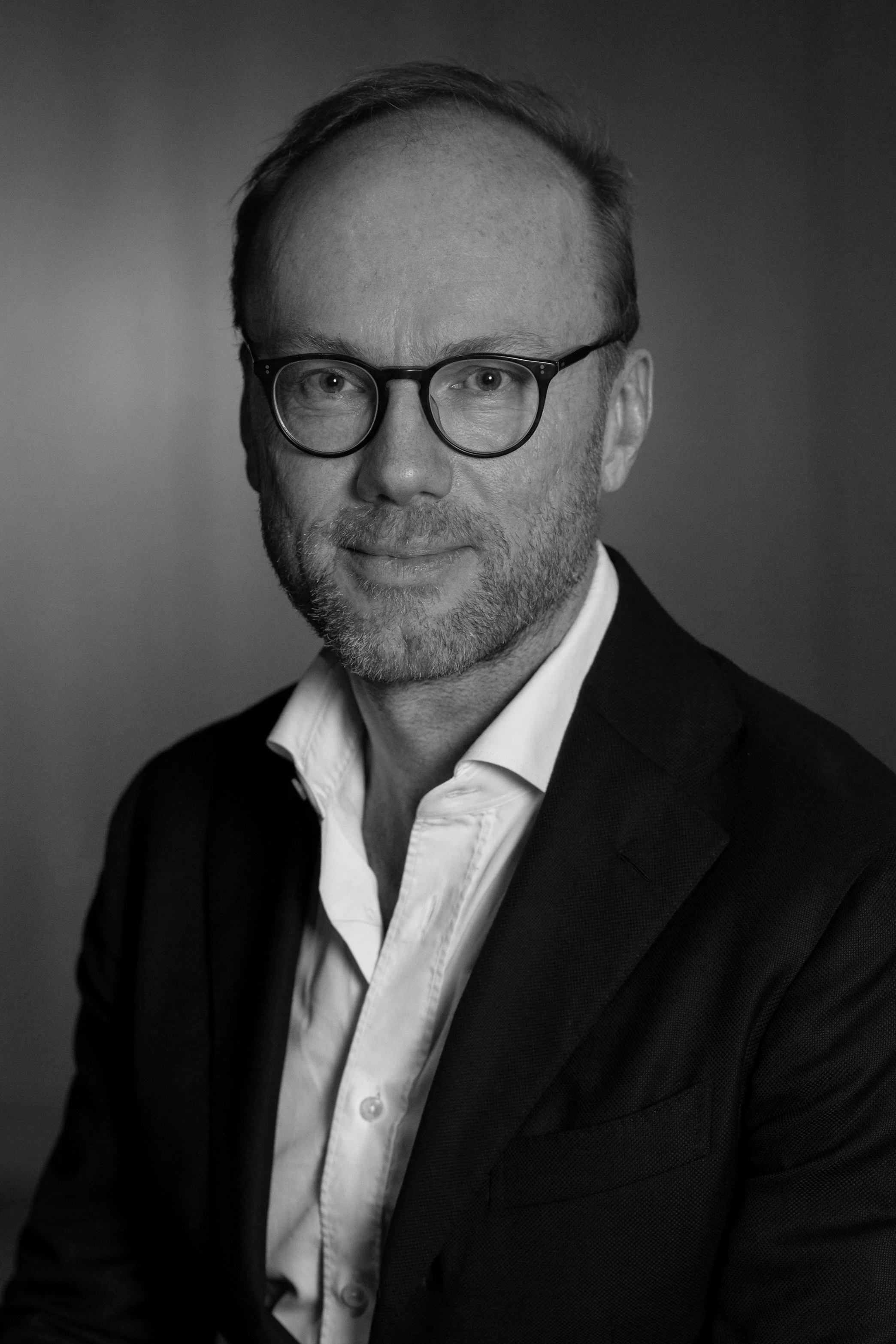 A middle-aged man with short hair and a beard, wearing a suit and tie, standing against a gray background.