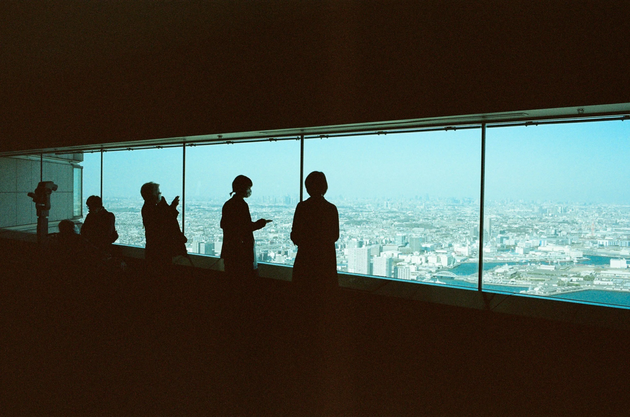 Silhouettes of people standing and sitting near a large window in a high-rise building, with a city skyline visible in the background.