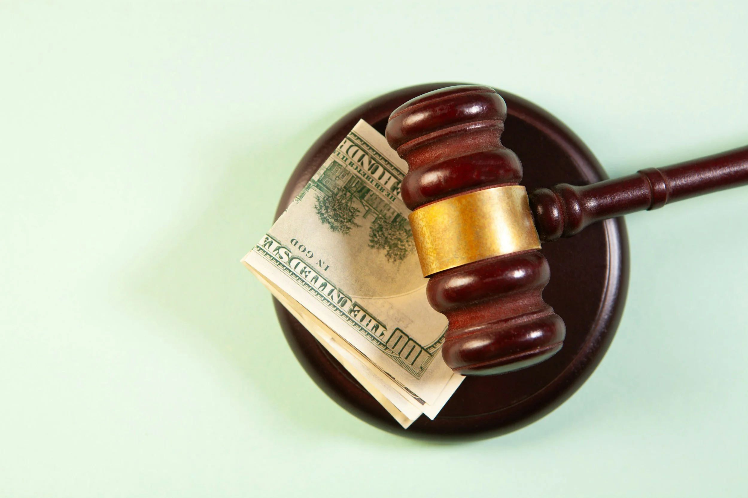 A wooden judge's gavel resting on a round base with a folded ten-dollar bill underneath, against a light green background.