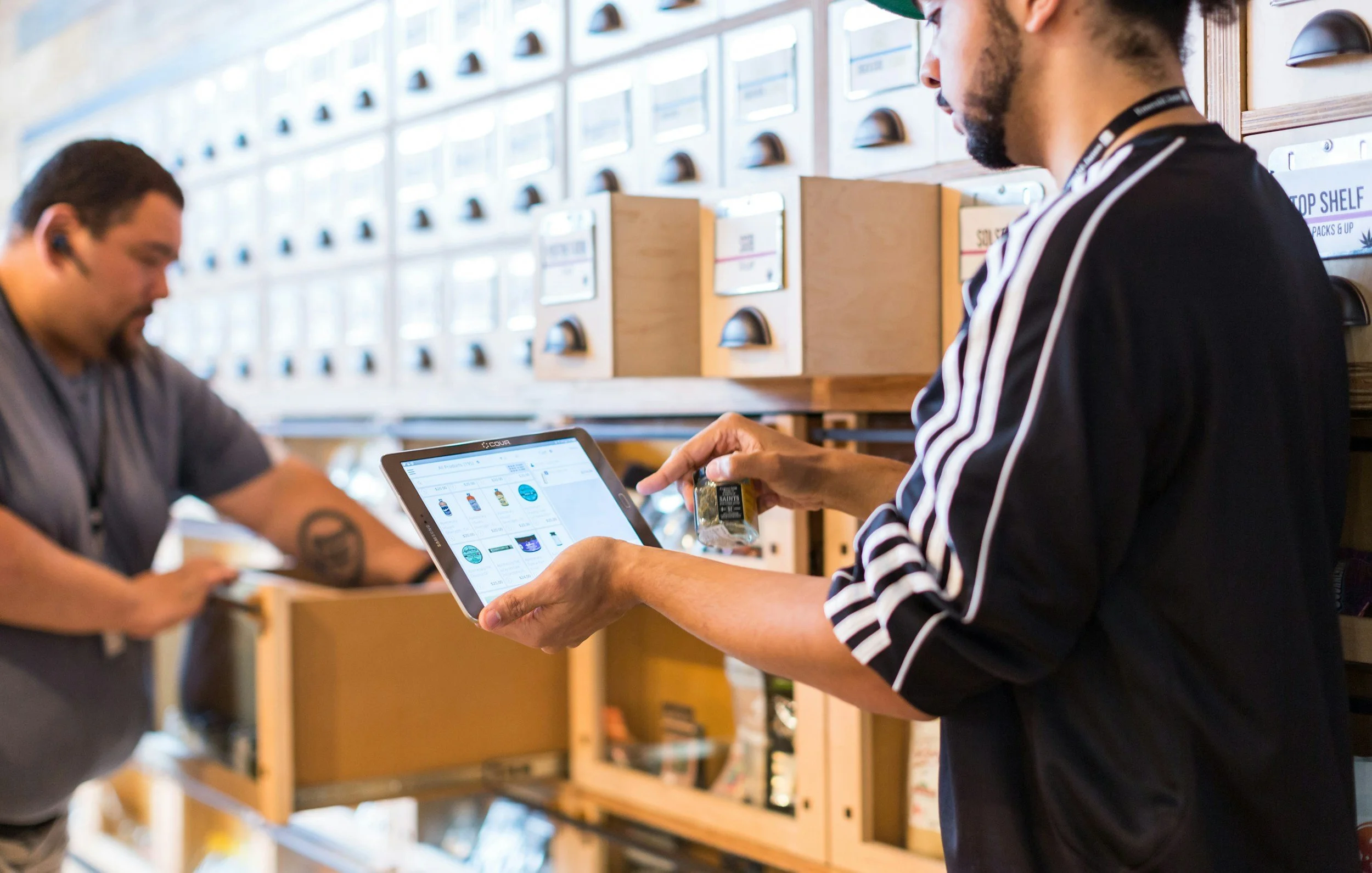 Two people at a retail store checkout counter, with one person holding a tablet displaying a digital shopping list or catalog, and the other person is holding a small product, both engaged in a purchase or transaction.